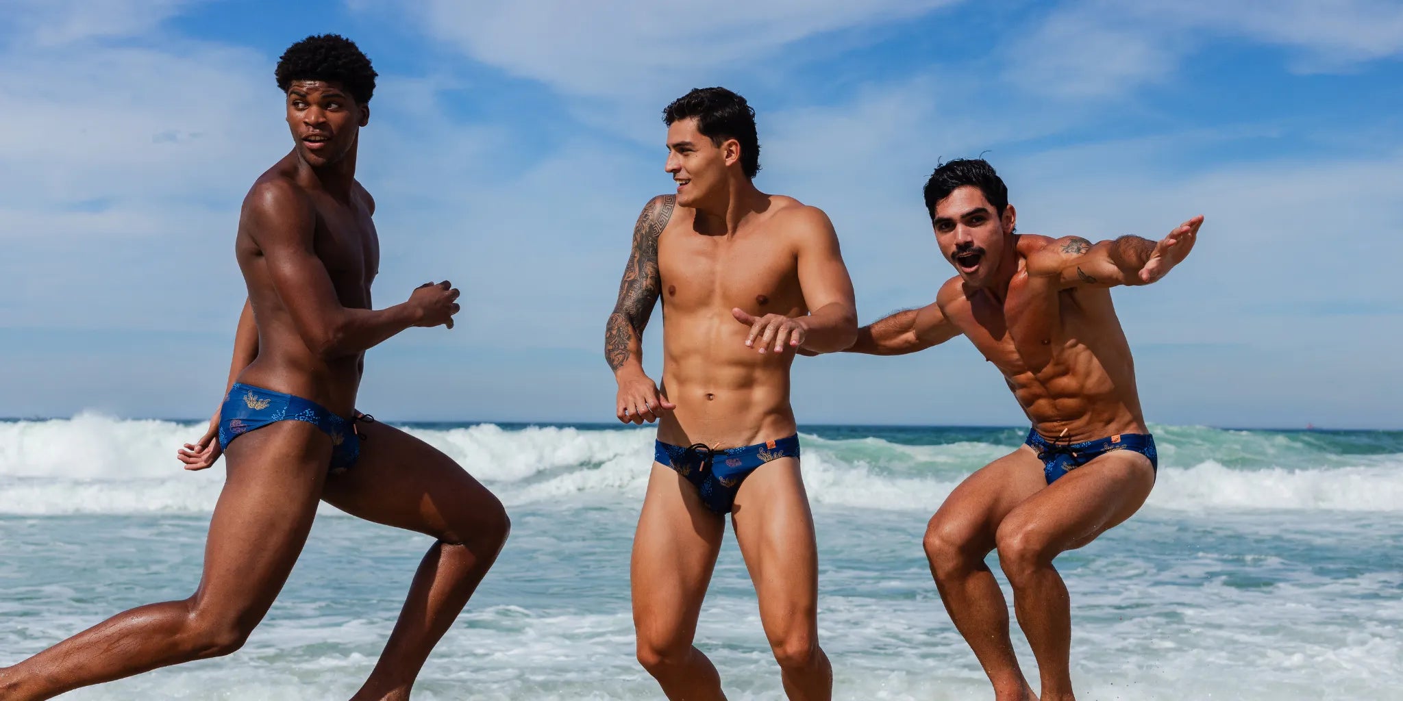 Three men in swim briefs running on a UK beach with ocean waves and sky in the background