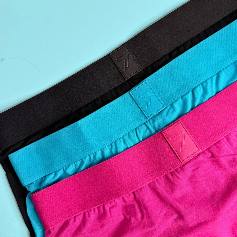 Three pairs of boxer briefs laid flat diagonally, showing the best elastic waistbands in black, bright turquoise, and vivid pink. Each waistband has a small square embossed 7 logo patch.