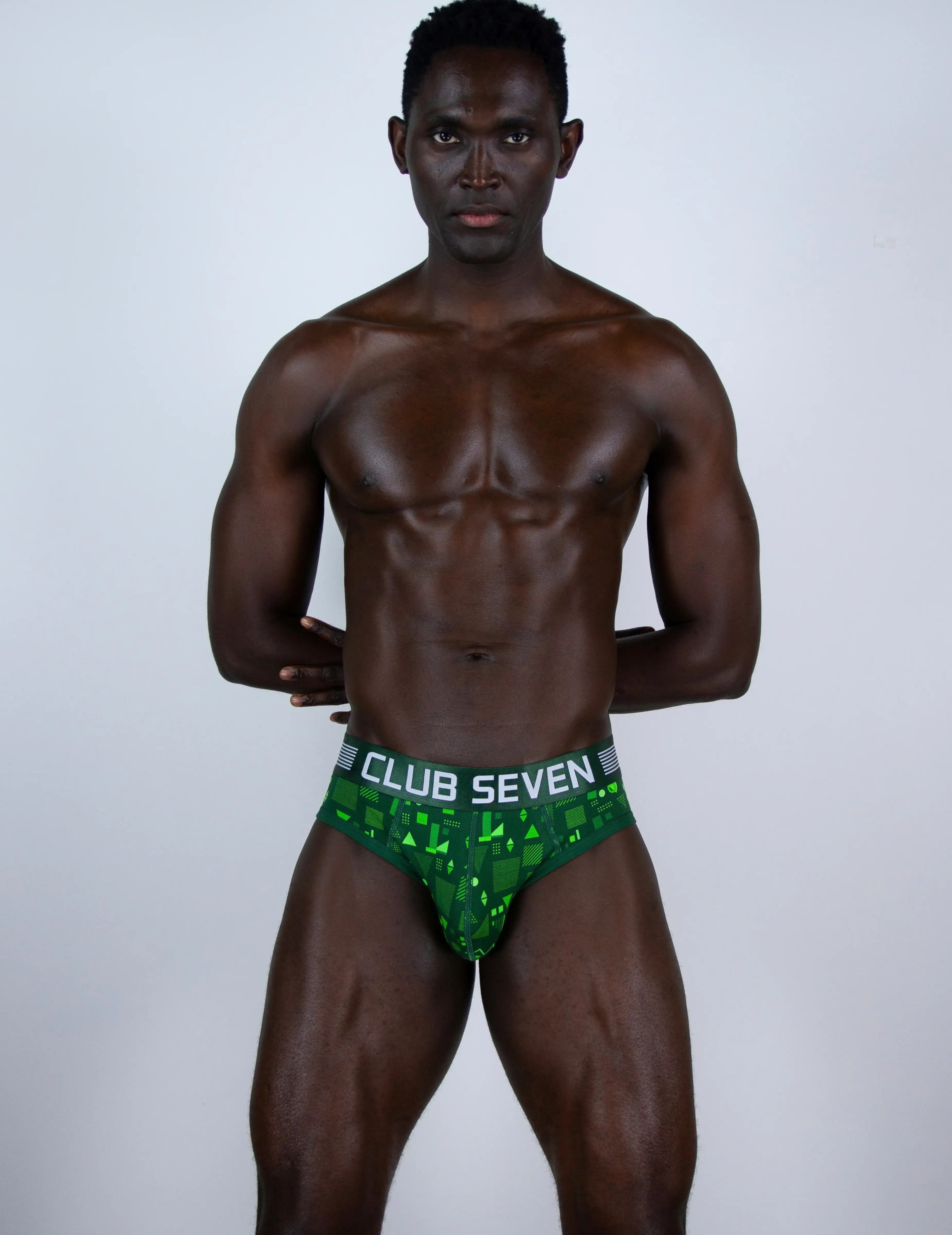 A muscular man modeling green patterned briefs with a white waistband that reads 'CLUB SEVEN' against a light gray studio background.