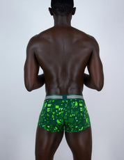 A man viewed from behind wearing green patterned boxer briefs with a gray waistband, set against a plain light gray studio background.