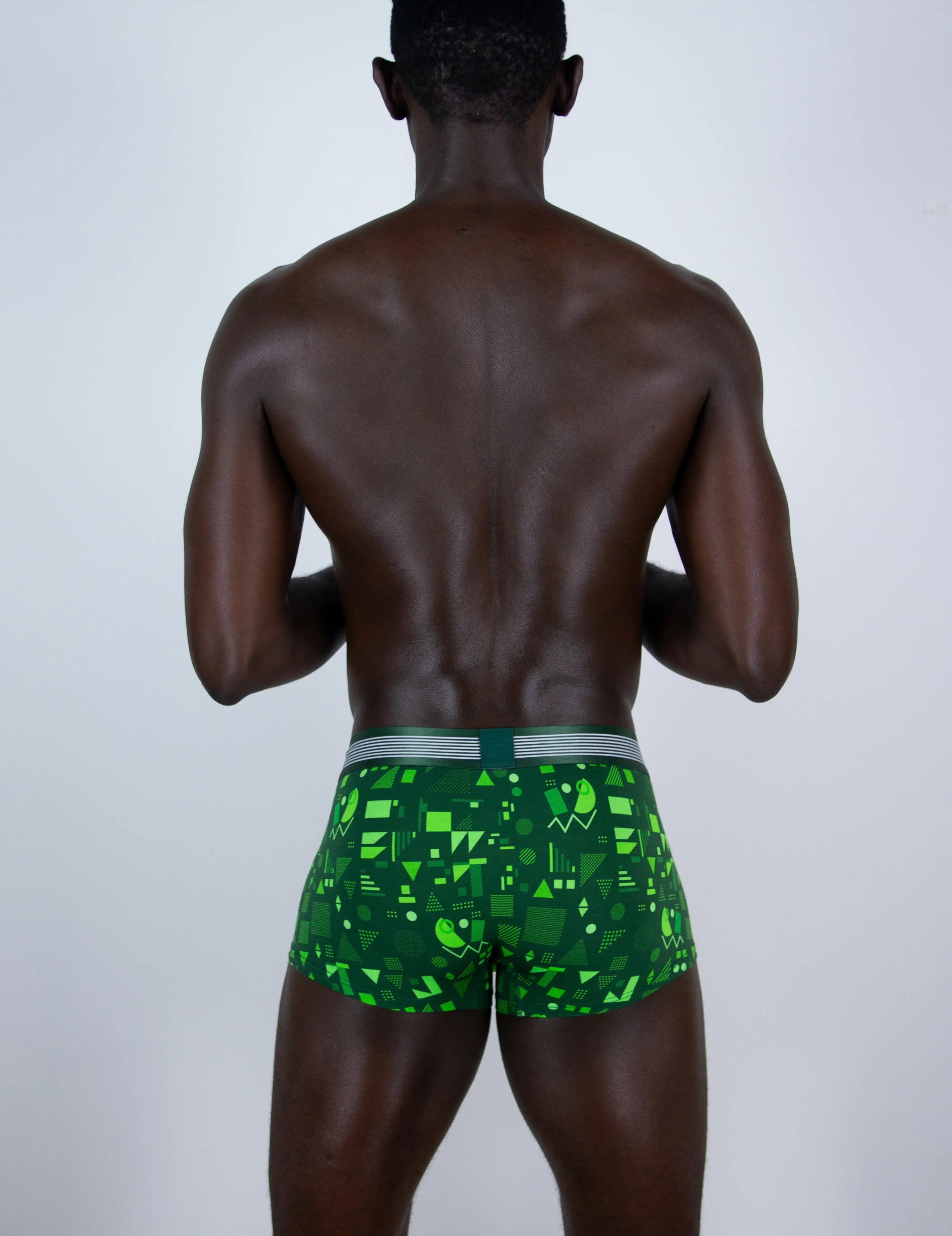 A man viewed from behind wearing green patterned boxer briefs with a gray waistband, set against a plain light gray studio background.