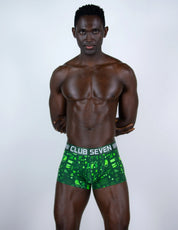 A shirtless man modeling green boxer briefs with a black and white pattern and a visible waistband that reads 'CLUB SEVEN'.