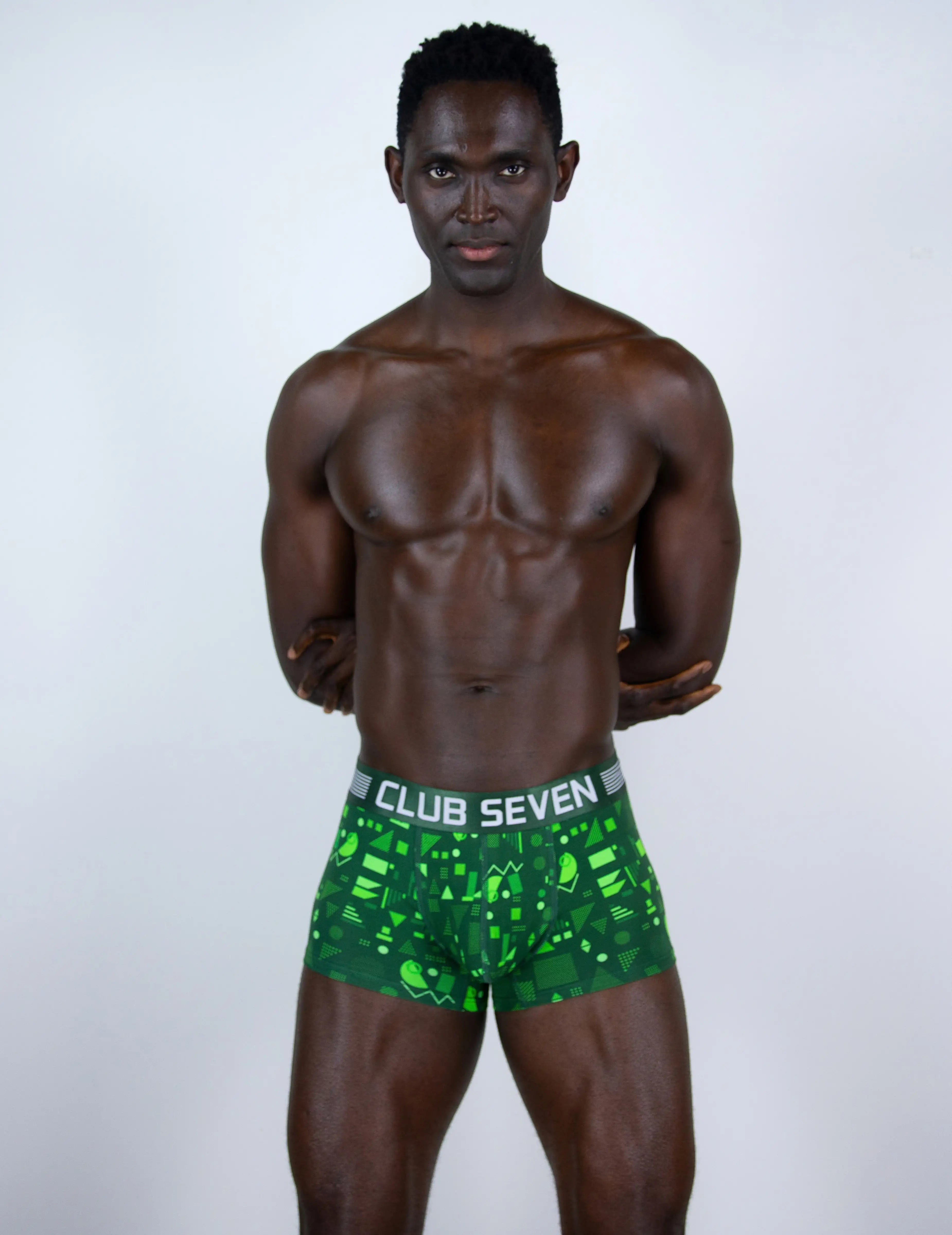 A shirtless man modeling green boxer briefs with a black and white pattern and a visible waistband that reads 'CLUB SEVEN'.