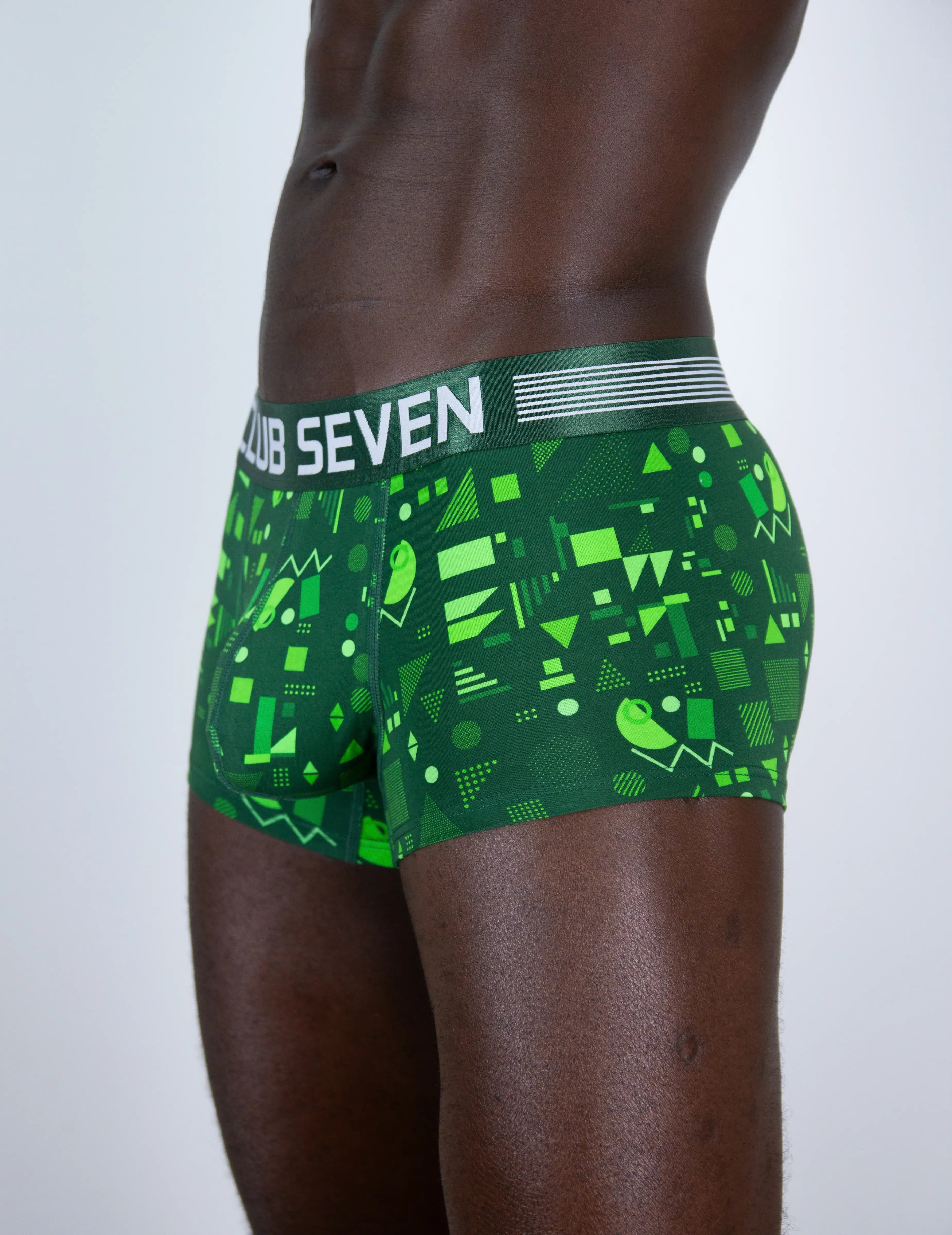 A close-up side view of a male model wearing green boxer briefs with a geometric pattern and a white waistband that reads 'CLUB SEVEN'.