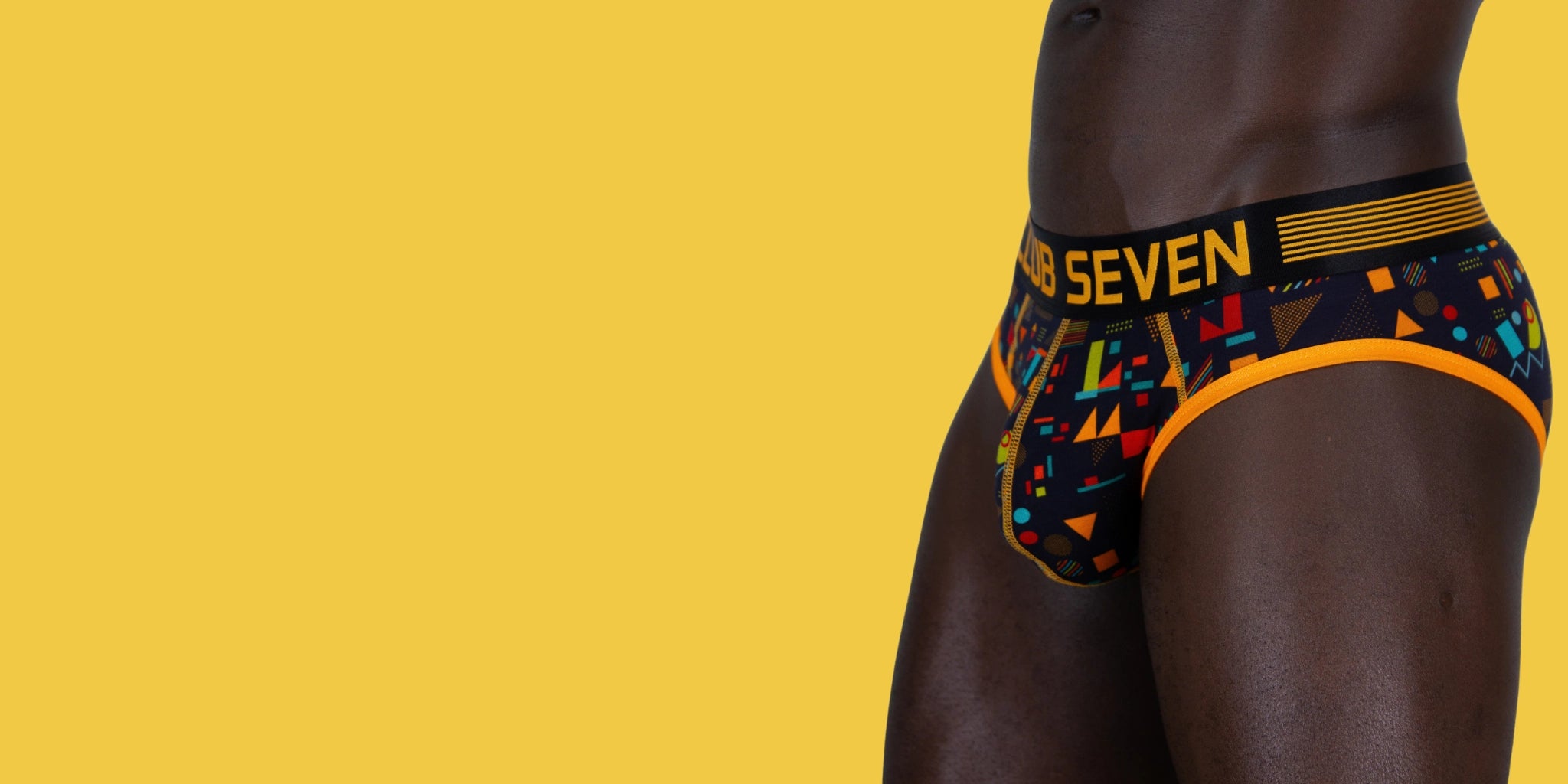A close-up side view of a model wearing patterned briefs with a big pouch, black waistband and multicolor geometric print, set against a solid yellow background.