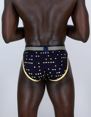 Rear view of a man wearing navy blue tanga briefs with a gold geometric pattern, a gold waistband, and a navy blue center tab, set against a plain light gray background.