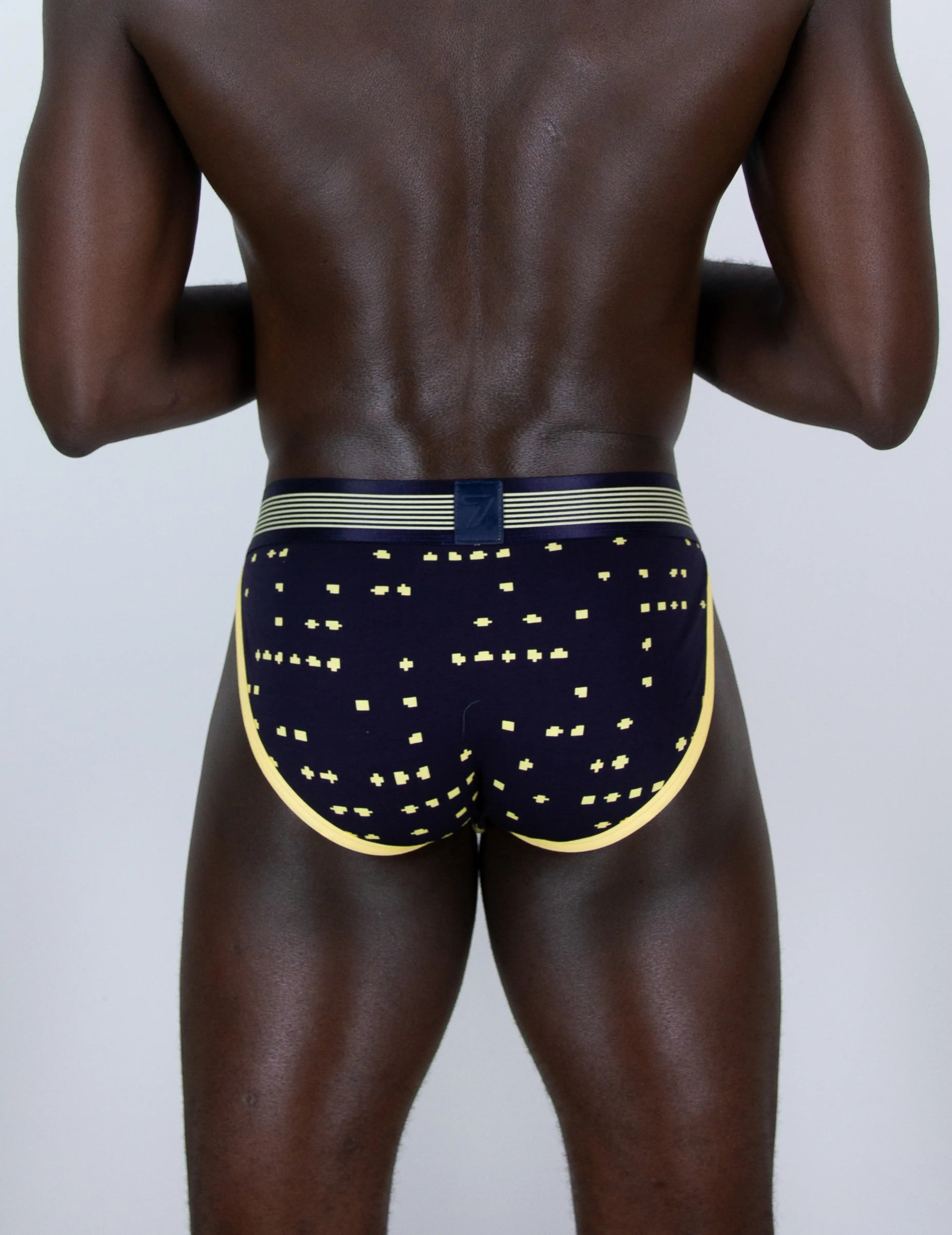 Rear view of a man wearing navy blue tanga briefs with a gold geometric pattern, a gold waistband, and a navy blue center tab, set against a plain light gray background.