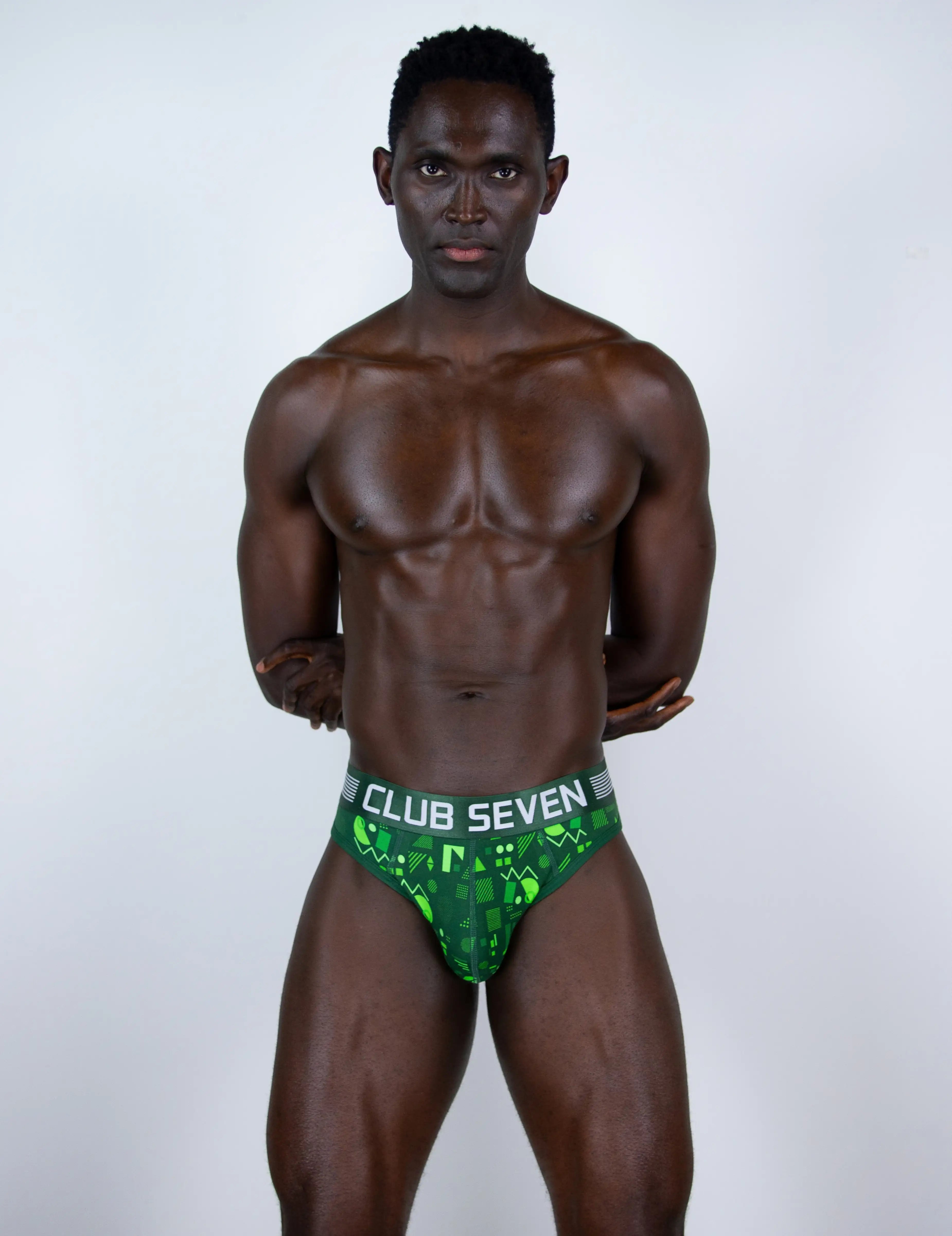 A muscular man modelling green-and-black patterned thongs with a big pouch, waistband that reads 'CLUB SEVEN' in white text, photographed against a plain light gray studio background.