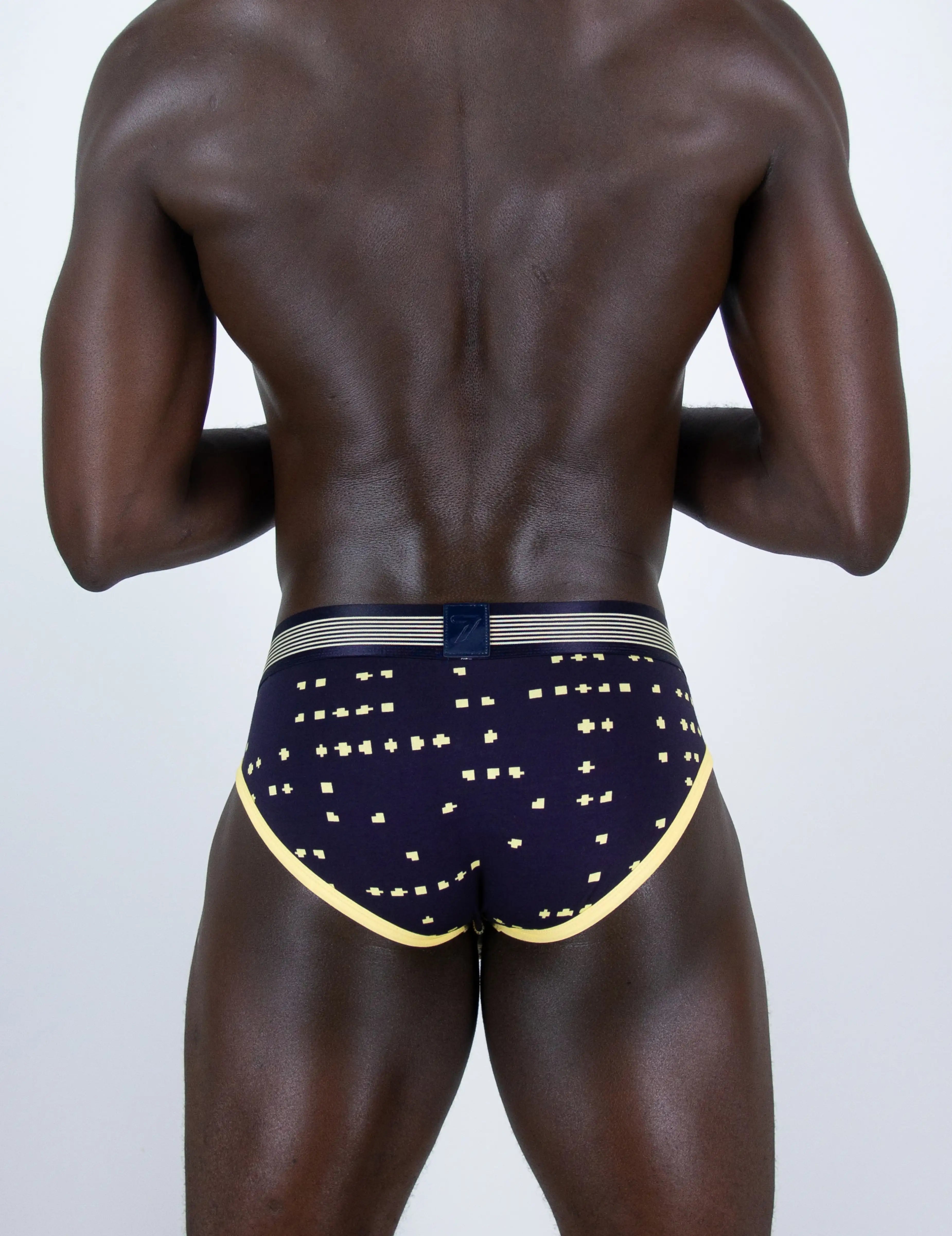 Rear view of a muscular man wearing navy blue boxer briefs with gold star patterns and a gold trim, on a light gray background.