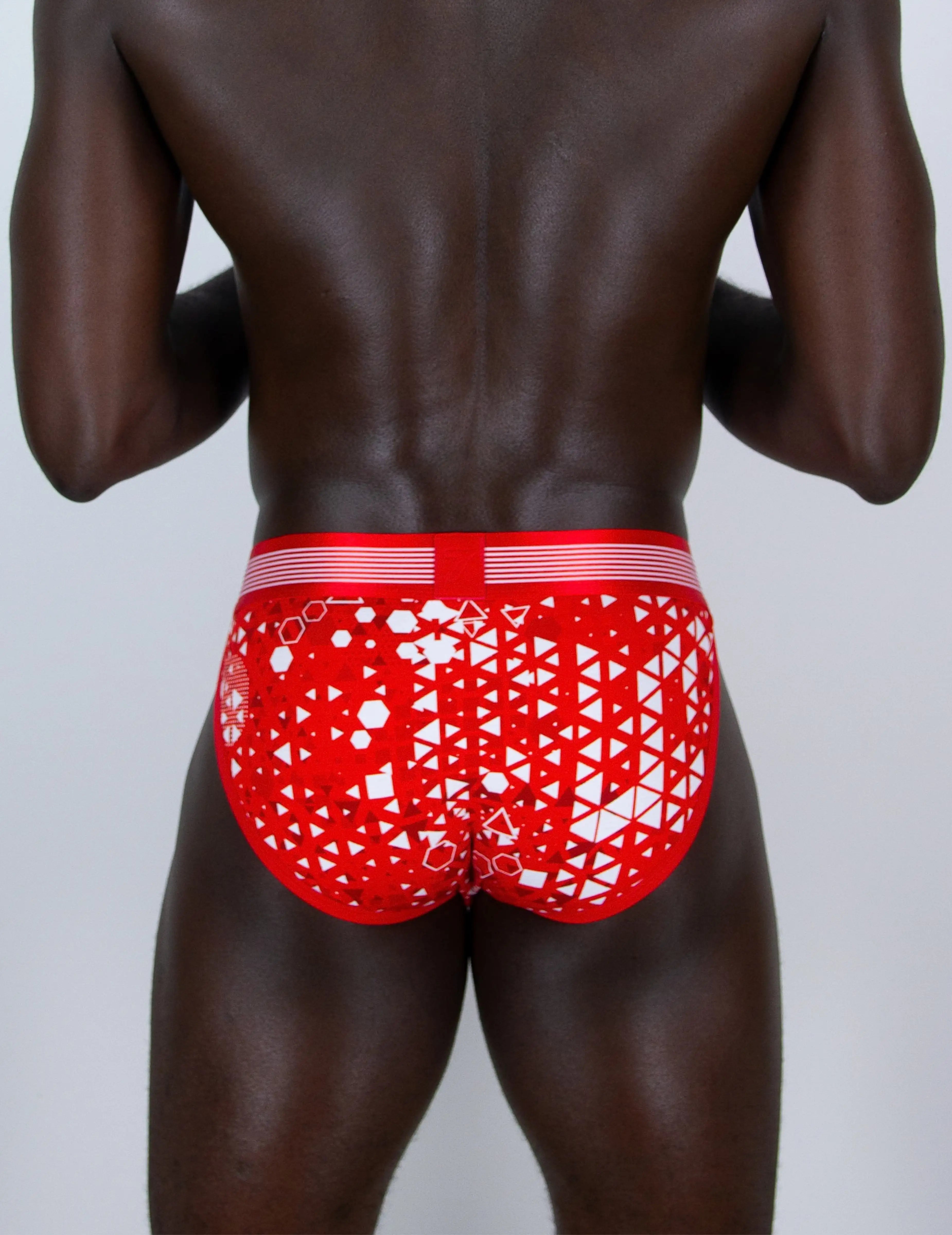 Rear view of a man wearing red and white patterned sports briefs against a light gray studio background.