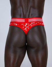 Rear view of a muscular male model wearing Christmas red-and-white patterned men's thongs with a big pouch against a light gray studio background.