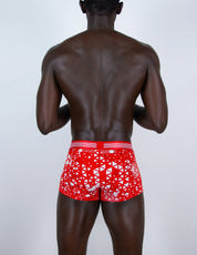 A man viewed from behind wearing red boxer briefs with a white geometric pattern and a red waistband, set against a plain light gray background.