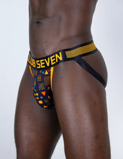 A close-up of a model's lower torso wearing men's low-rise jockstraps with a big pouch. The underwear features a black waistband with yellow stripes and 'CLUB SEVEN' printed in yellow, and a geometric multicolor pattern on the front pouch.