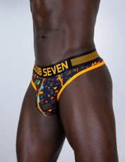 A close-up of a male model wearing a low-rise men thong with a big pouch, a black, orange, and multicolor geometric pattern. The waistband features the text 'CLUB SEVEN' in yellow on a black background.