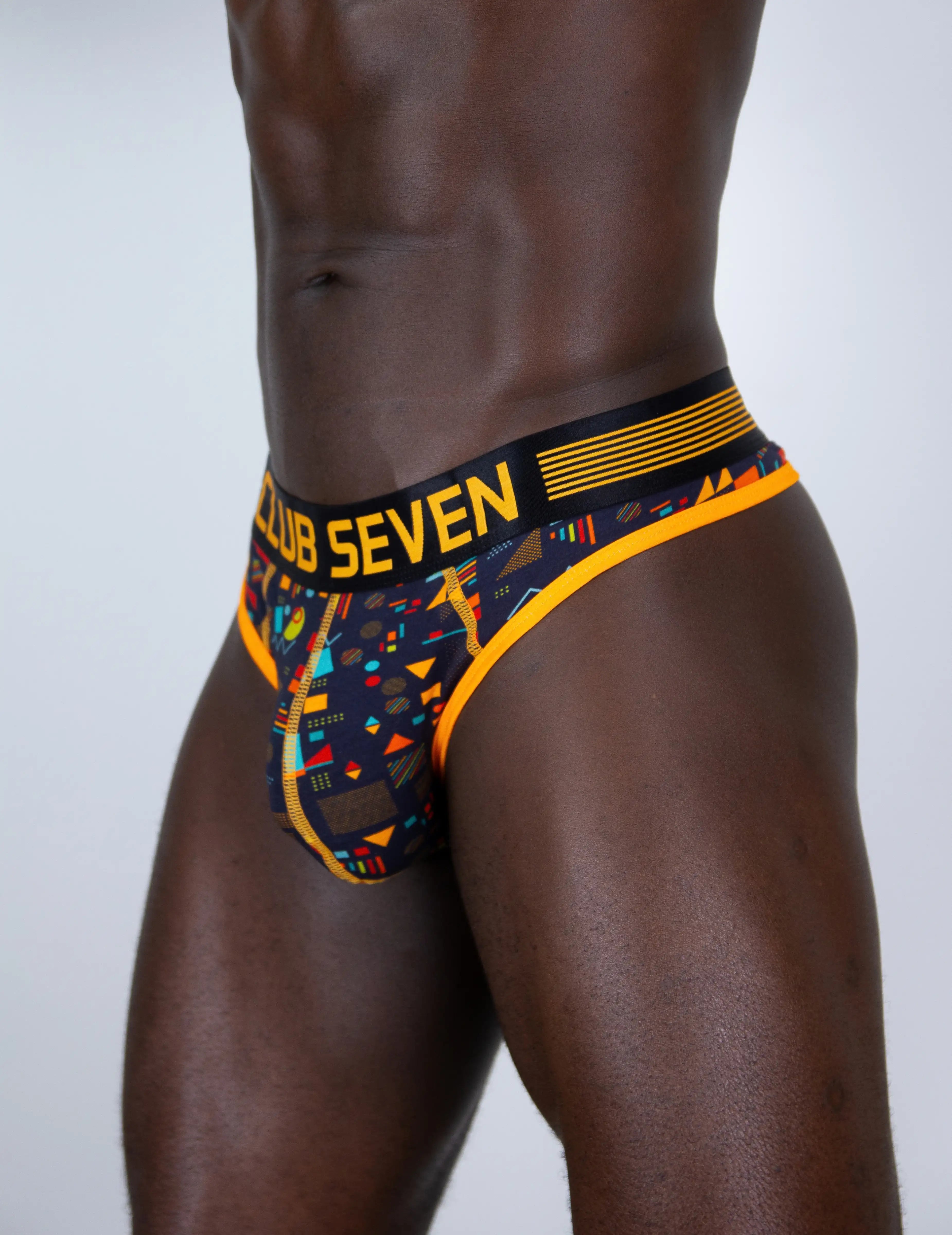 A close-up of a male model wearing a low-rise men thong with a big pouch, a black, orange, and multicolor geometric pattern. The waistband features the text 'CLUB SEVEN' in yellow on a black background.