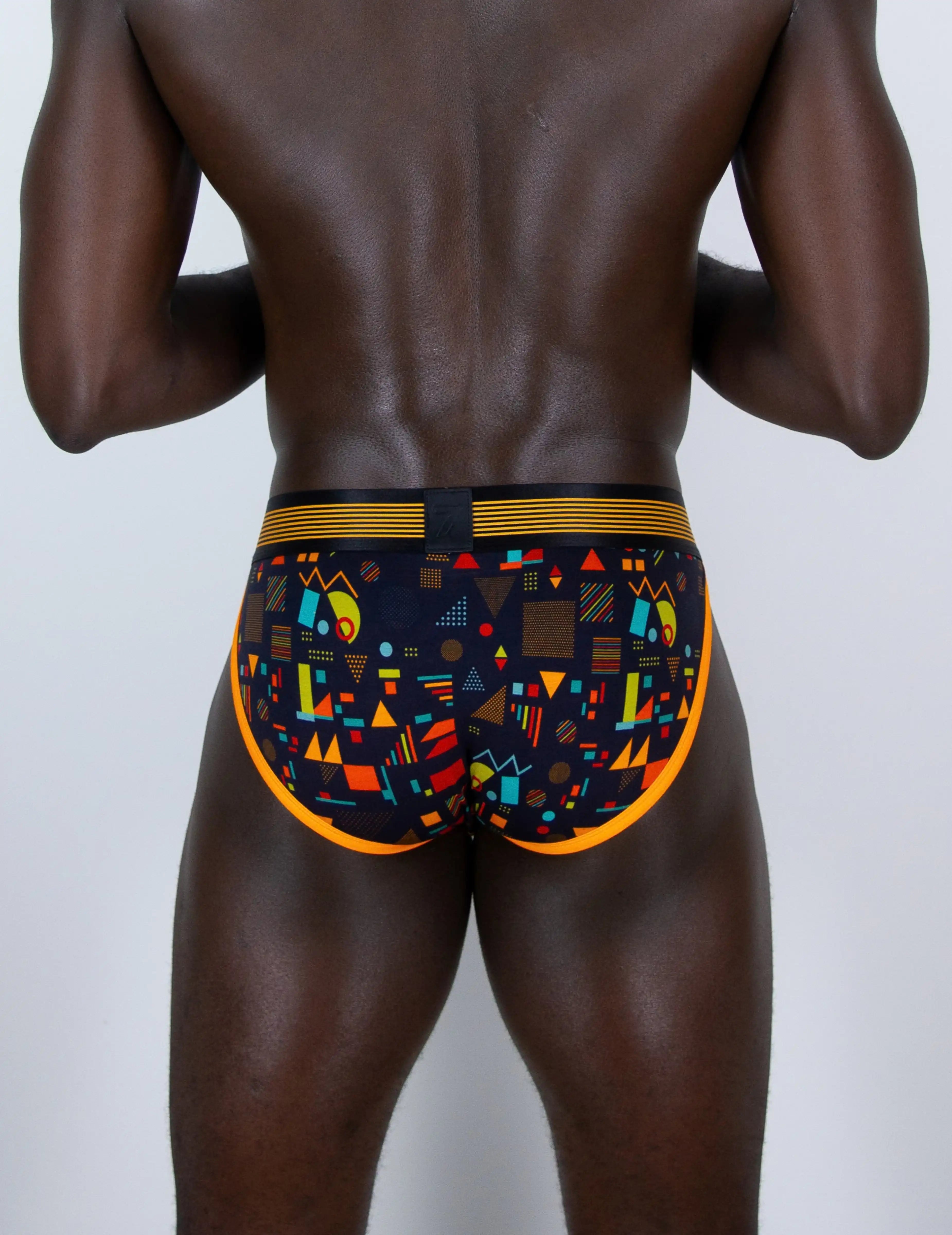 Rear view of a male model wearing low-rise tanga sports briefs with a colorful geometric pattern on a black background, featuring a gold waistband and orange side trim.