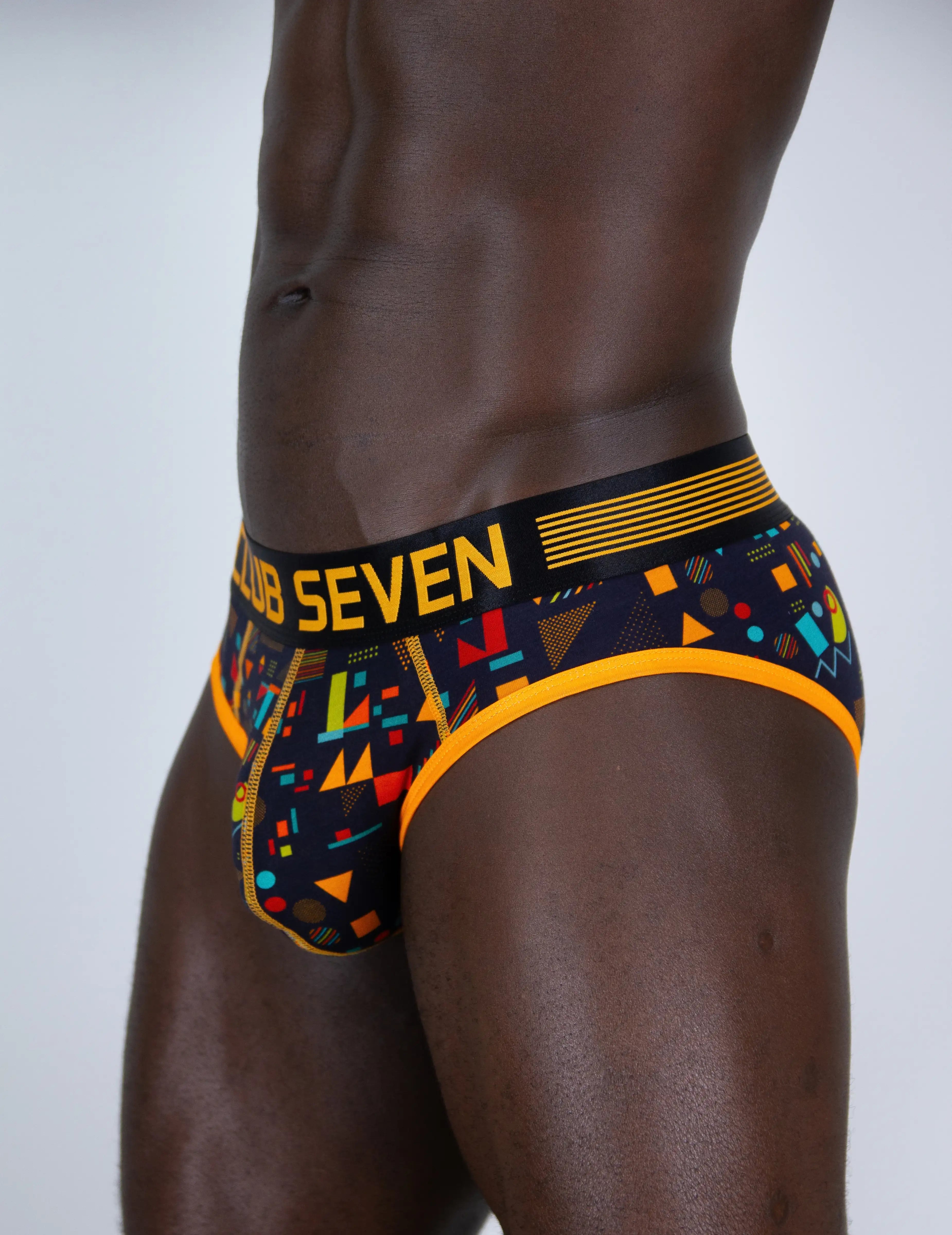 A close-up of a man wearing Club Seven branded men's briefs. The briefs feature a black base with a vibrant multicolour geometric pattern (yellow, orange, red, blue, green) and a black waistband with yellow text and stripes.