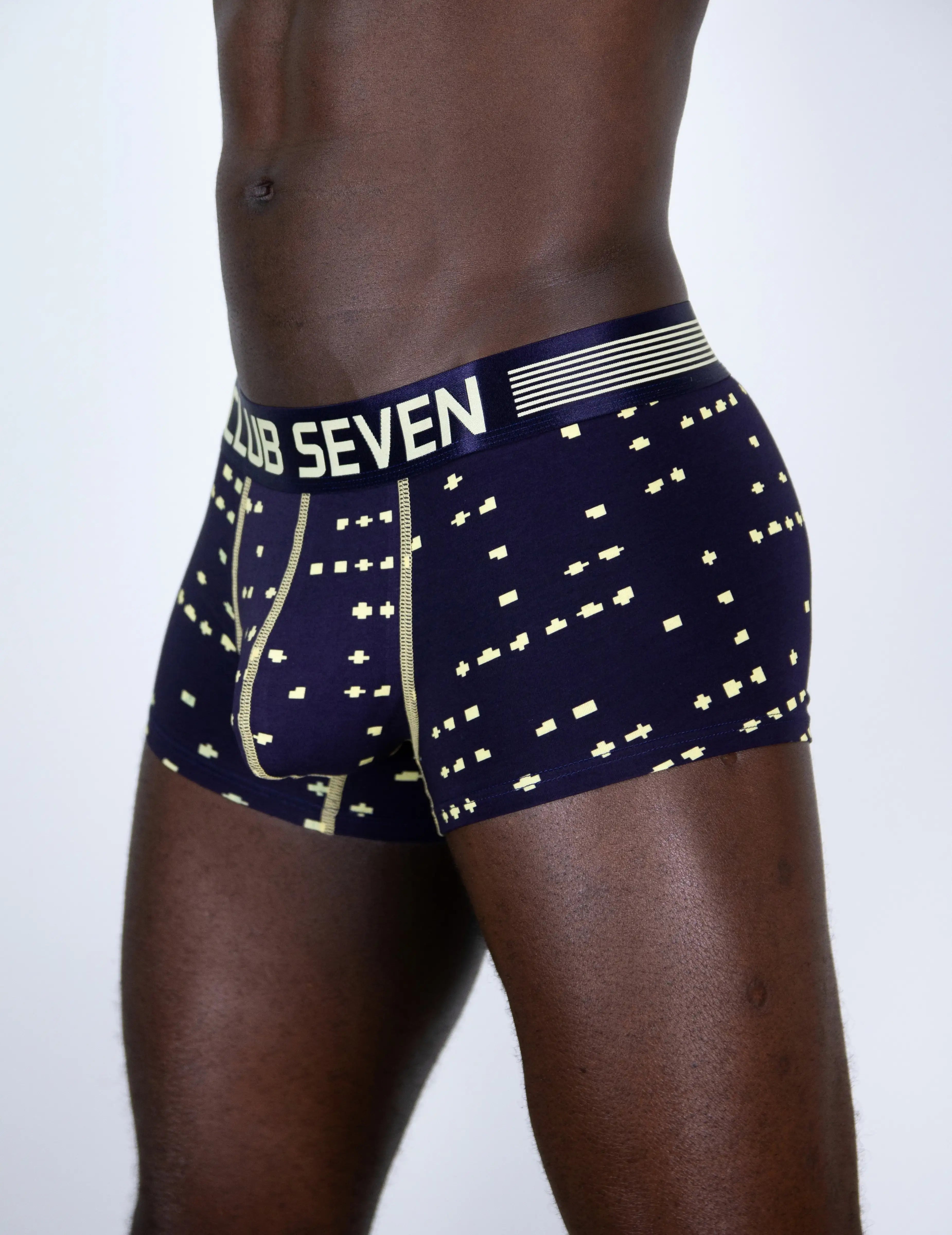 A close-up of a male model's lower torso wearing Club Seven boxer briefs. The underwear is navy blue with a gold geometric pattern and a matching navy waistband featuring the white 'CLUB SEVEN' brand name.